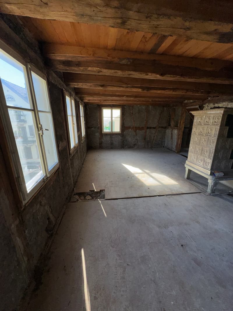 Empty room with unfinished walls and exposed wooden beams, several large windows, one window has a metal protective bar.
