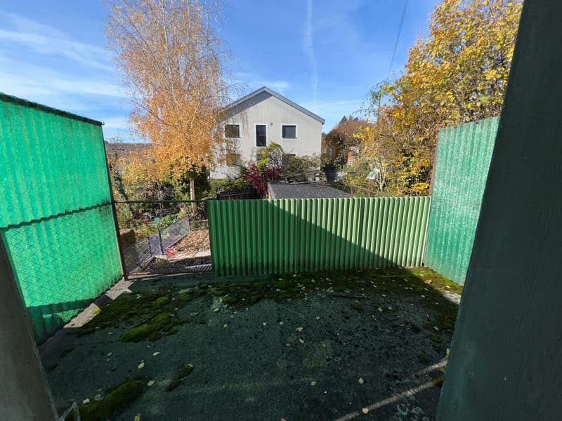 Outdoor space with green fence, house and greenery in background, paved ground