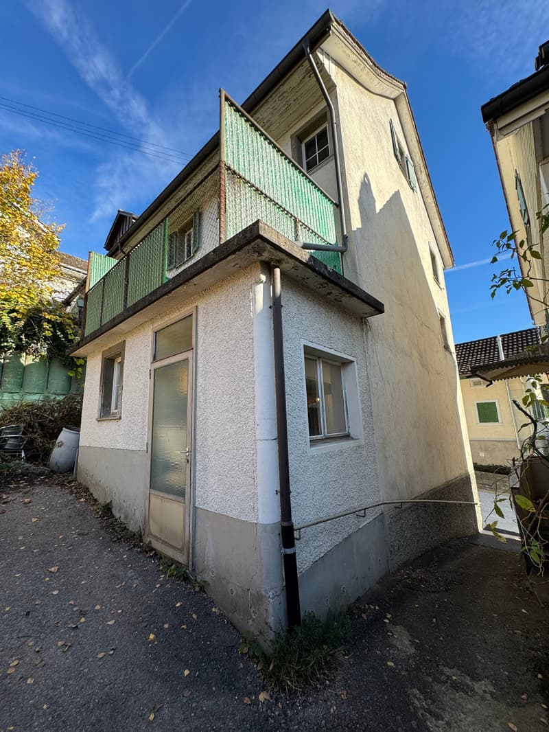 Two-story house, concrete wall, glass balcony railing, windows, entrance door, plants, sidewalk