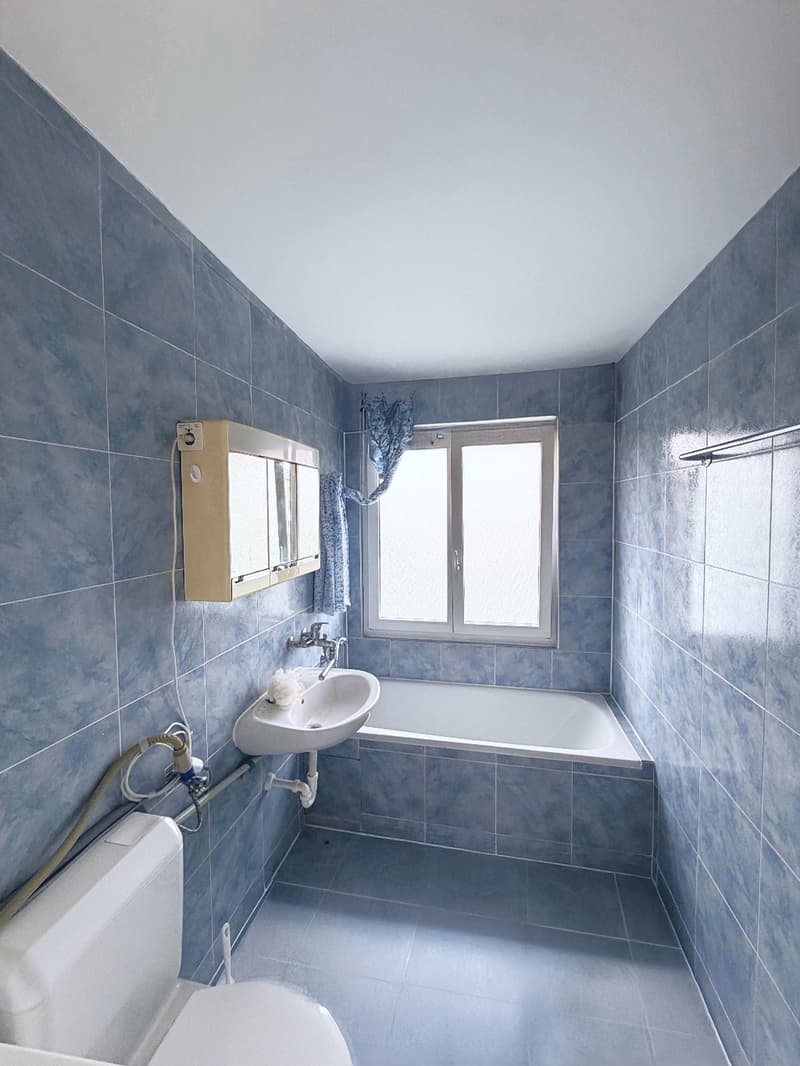Bathroom with blue tiles, white bathtub, white sink, white toilet, window with blue curtain, mirror with cabinet