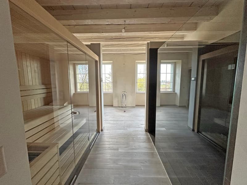 This image shows the interior of a room with a sauna. The room has wooden beams on the ceiling, large windows, and a glass-enclosed sauna area. The floor is made of wood planks. The space appears to be in a state of renovation or construction, with an ope