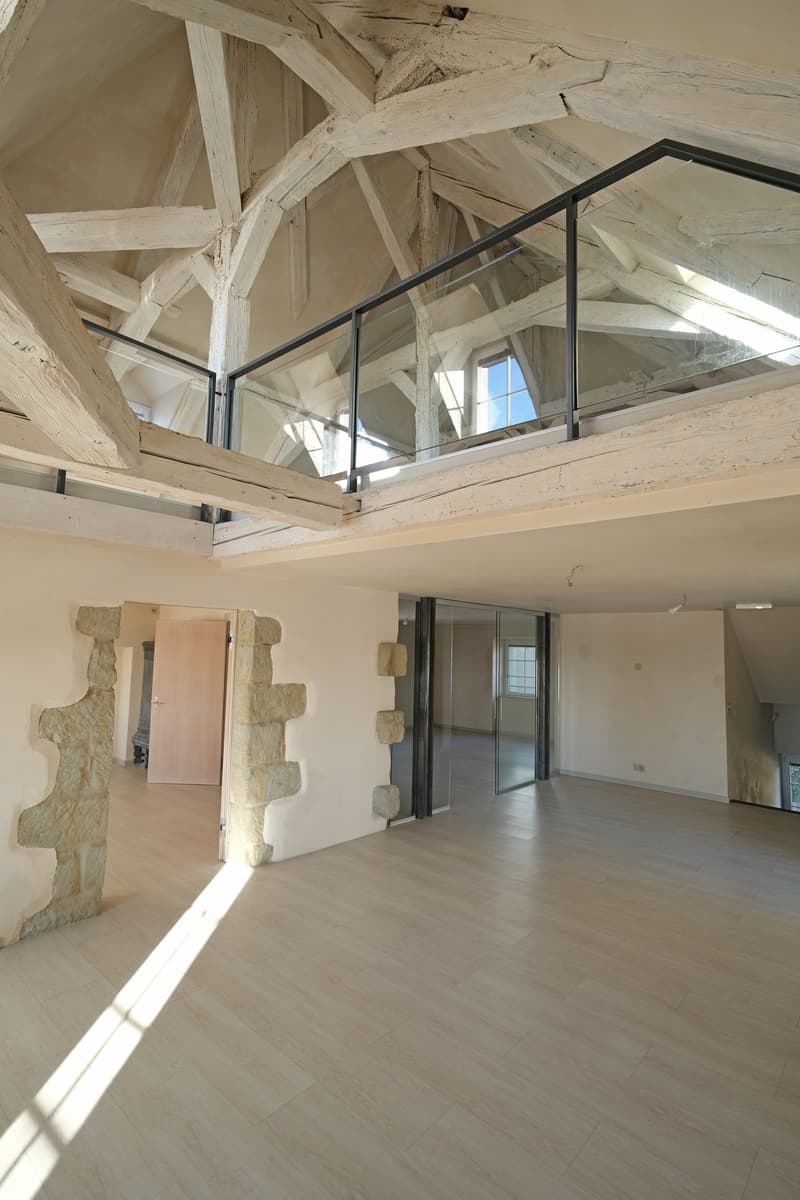 Large open-concept space with exposed wooden beams and trusses in the ceiling, stone walls, and hardwood flooring. The space features a mezzanine-style balcony or loft area overlooking the main living space.