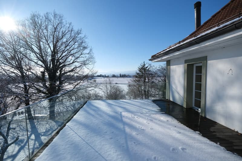 2-story house with a balcony/terrace overlooking a snowy landscape, bare trees in the foreground, and a lake or body of water in the background.