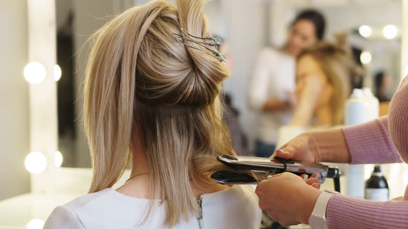 A person with blonde hair getting their hair styled in a salon, a woman holding a straightening iron in front of them, a blurry view of a person standing behind them with bottles in front of them, and a mirror with a light on the side.