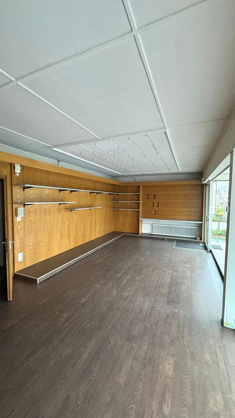 Empty room with dark wood flooring, wooden wall with shelves, an exit to the left side, a large window and sliding doors to the right side, white ceiling, white radiator under the window.