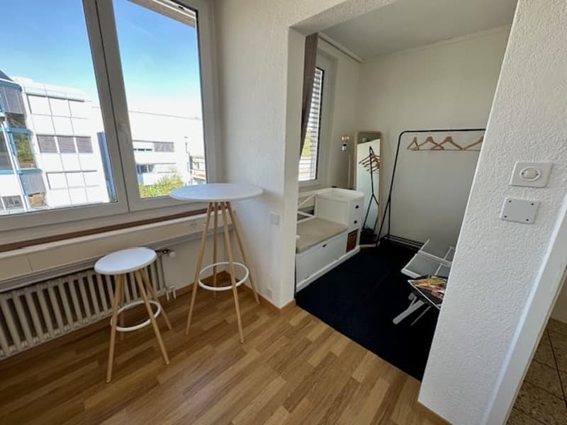 Small room with wooden floor, white walls, large window, round table, stools, radiator, built-in bench, mirror, clothes rack, and rug.