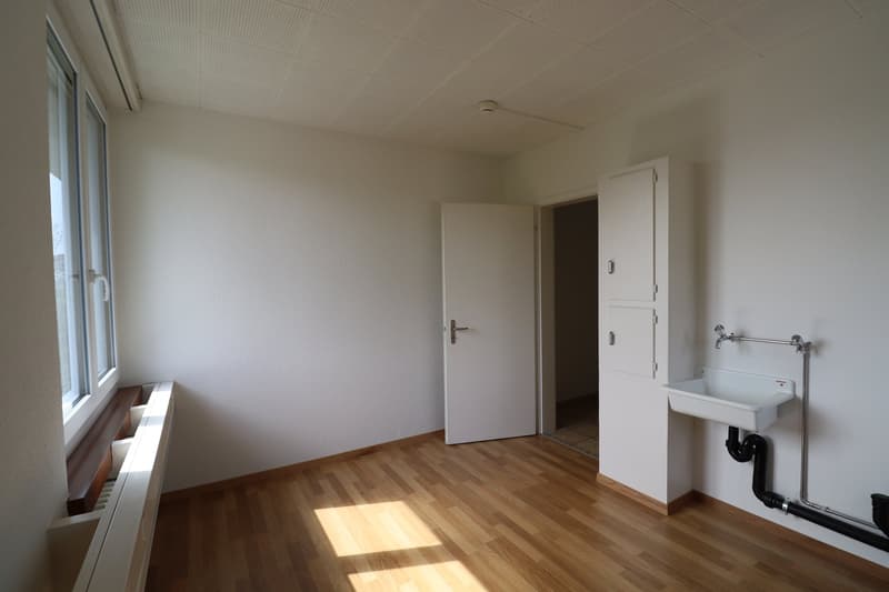 Empty room with white walls, wooden floor, a window, a door, a sink with a pipe and radiator below the window.