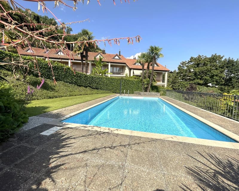 The image shows a residential complex with a swimming pool surrounded by lush greenery and palm trees. The buildings have a Mediterranean-style architecture with red tile roofs. The pool is rectangular in shape and appears to be well-maintained.