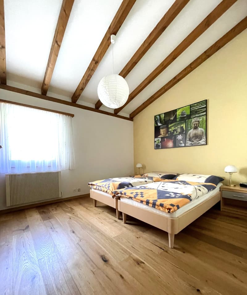 The image shows a bedroom with a vaulted ceiling featuring exposed wooden beams. The room has hardwood flooring and a large window that provides natural light. The bed is adorned with a colorful patterned comforter, and there is a framed artwork depicting
