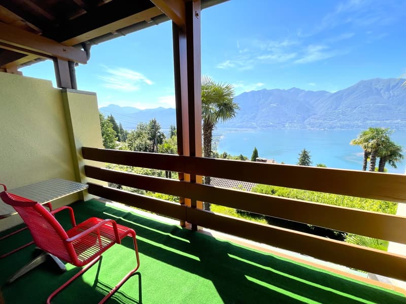 Wooden balcony with a scenic view of the lake and mountains, surrounded by palm trees and lush greenery. The balcony has a green artificial grass floor and red metal chairs, providing a comfortable outdoor space to enjoy the beautiful natural surroundings