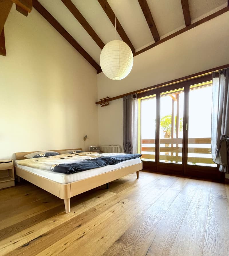 Spacious bedroom with wooden beams on the ceiling, hardwood flooring, large windows with sliding glass doors leading to a balcony or terrace, a white paper lantern light fixture, and a wooden bed frame with a blue bedspread.