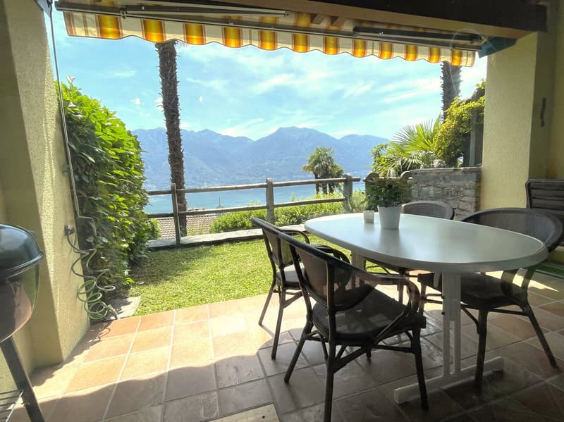 Covered outdoor patio with a round table and chairs, overlooking a scenic view of mountains and greenery. The patio has a retractable awning and is surrounded by lush vegetation.