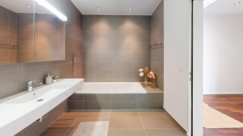 Modern bathroom with white tiles, free-standing bathtub, under-mount sink, and a large mirror. Small potted plants are on the sink, and there is a rug on the floor.