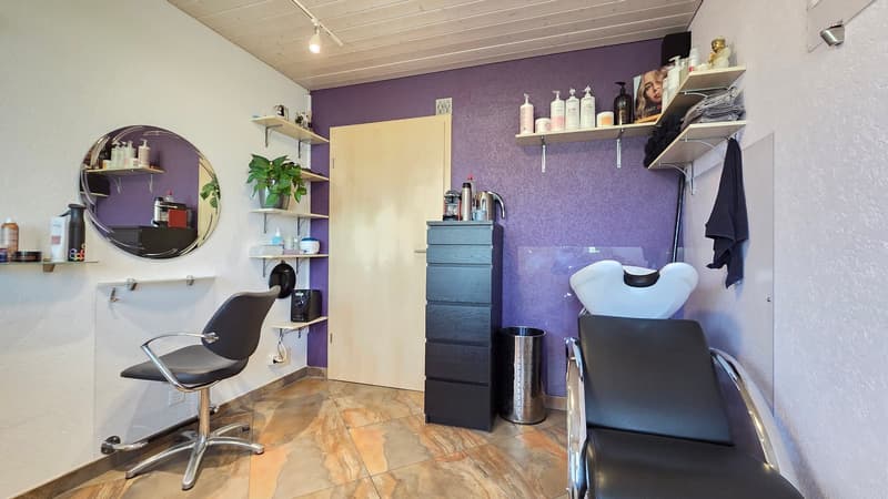 A salon room with purple walls, tiled floor, two black chairs, a desk with drawers, a mirror, shelves with various items, a sink, and a trash can.