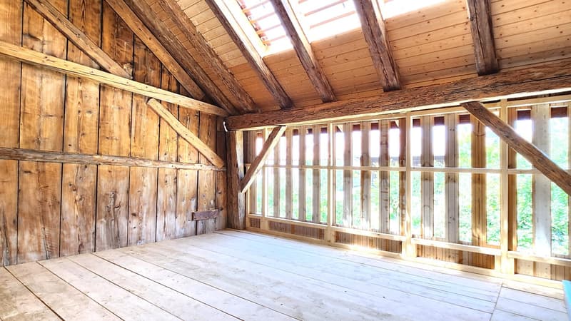 The image shows the interior of an old wooden barn. The walls are made of weathered wooden planks, and the ceiling has exposed wooden beams. The floor is also made of wooden boards. The barn has large windows that allow natural light to enter the space.