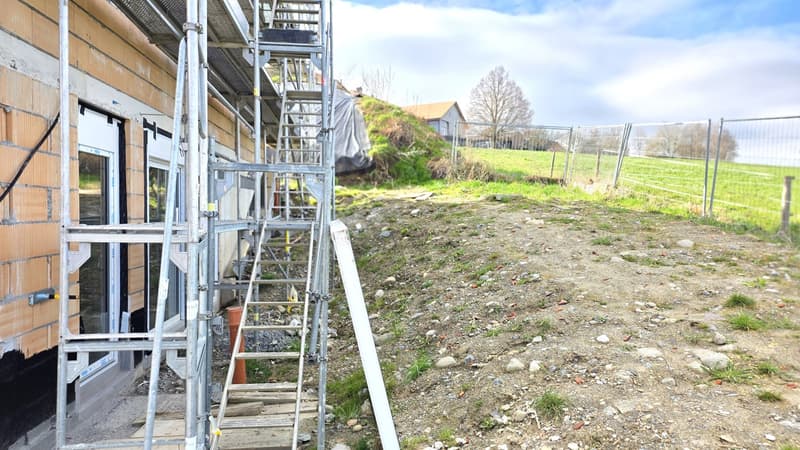 House under construction, brick wall, scaffolding, dirt ground, metal fence, grassy field, house in the distance