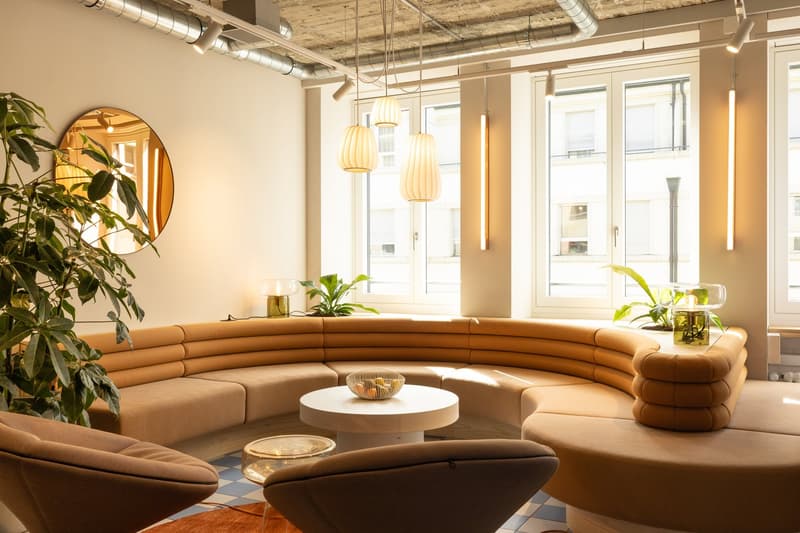 Spacious living room with curved beige sofa, round coffee table, potted plants, and pendant lighting fixtures. Large windows provide natural light and a view of the exterior.