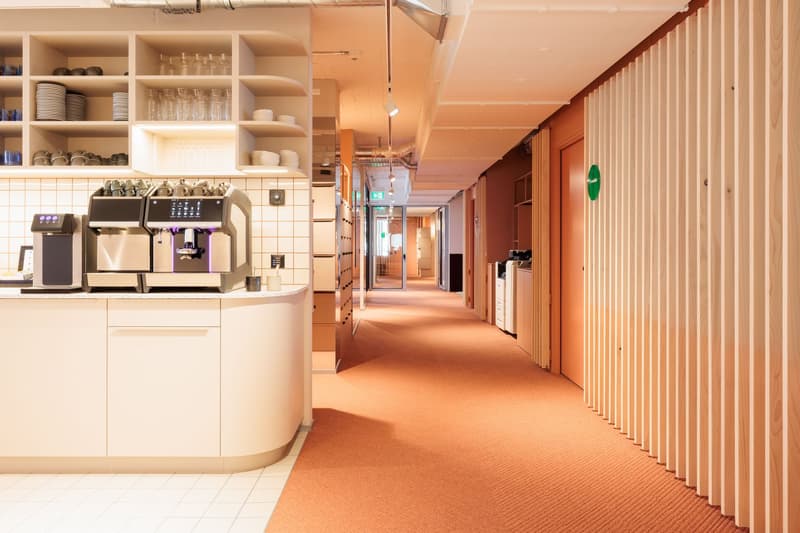 The image shows a modern office or commercial space with a kitchen area featuring a coffee machine and dishwasher. The space has a long hallway with wooden paneling and shelving units along the walls, indicating it is likely a shared workspace or office e