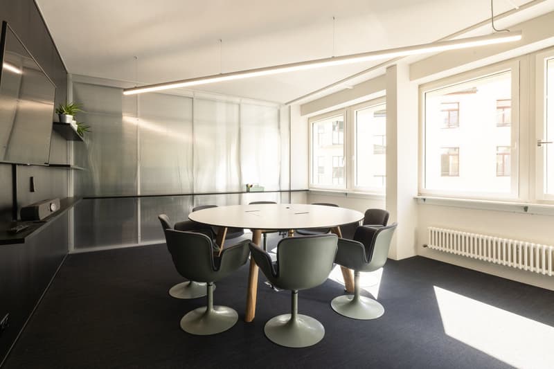 Modern office space with a round glass table, black swivel chairs, and large windows providing natural light. The room has a minimalist design with white walls, black flooring, and built-in storage units. A dishwasher is visible in the corner of the room.