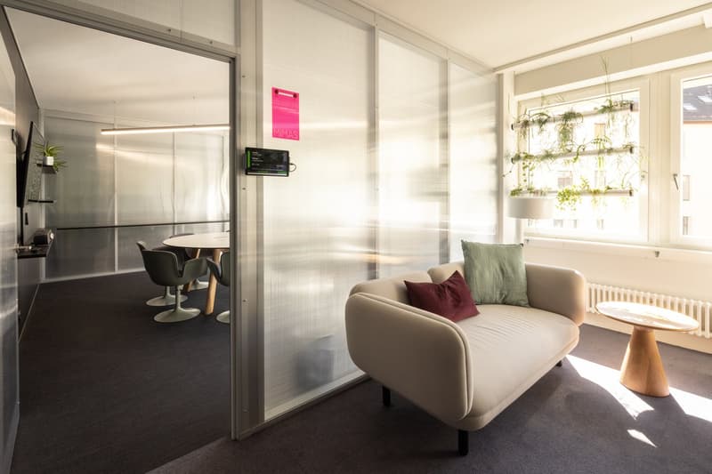 Modern office space with glass walls, comfortable seating, a dining table, and a hanging plant display. The space has an open and bright atmosphere.