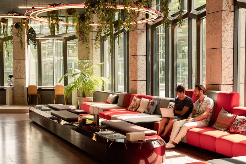 Spacious lobby area with large windows, red and gray sofas, coffee tables, potted plants, and hanging light fixtures.