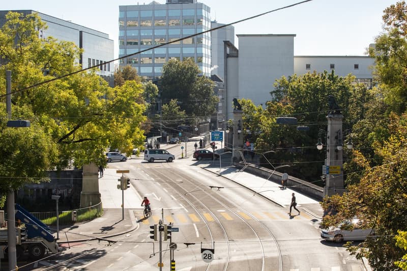 The image shows a city street with a tram line running through it. There are several multi-story buildings, some with glass facades, surrounded by trees with yellow foliage. There are a few pedestrians and vehicles visible on the street.