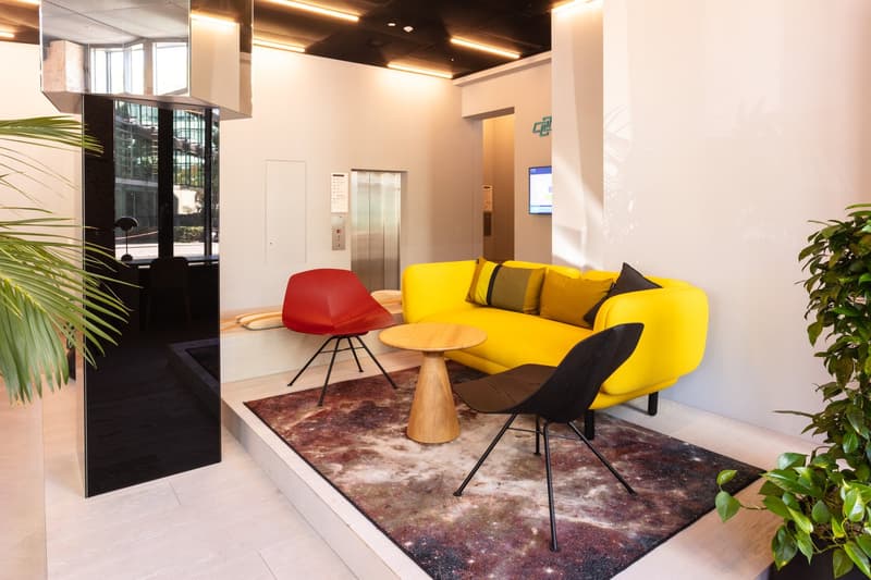 Spacious lobby with modern and colorful furniture, including a yellow sofa, red chair, and wooden coffee table. The room has large windows, a digital display, and potted plants, creating a bright and inviting atmosphere.