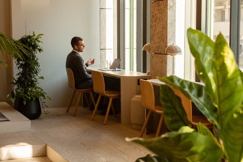 Modern office space with large windows, wooden desk and chairs, potted plants, and a laptop on the desk