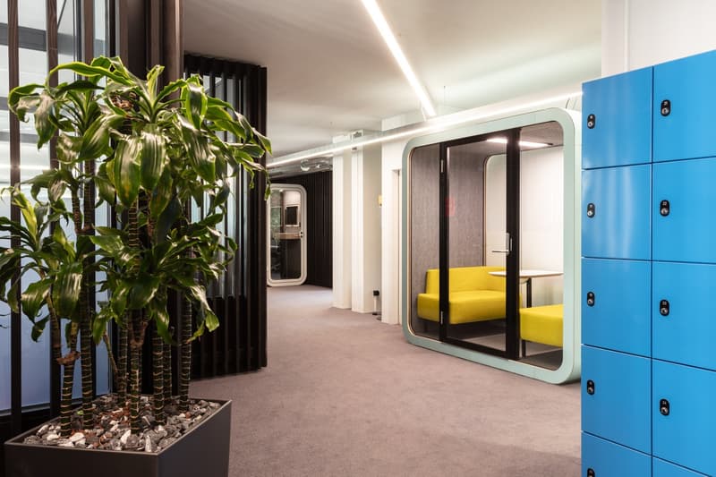 The image shows a modern and stylish office space with a large plant in the foreground, blue lockers on the left, and a yellow sofa in a glass-enclosed room in the background.