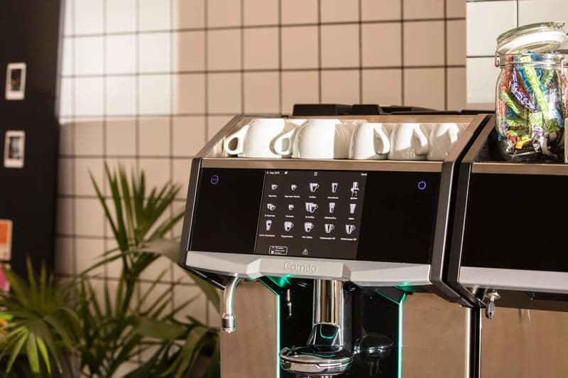 A modern coffee machine with a digital display showing various drink options. The machine has a sleek black and silver design and is placed on a kitchen counter next to a plant and a jar filled with colorful candies.