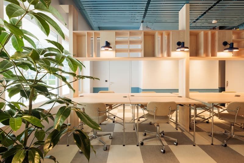 Open office space with large windows, wooden shelving units, and a conference table with chairs. The space has a modern, minimalist design with a blue ceiling and natural lighting.