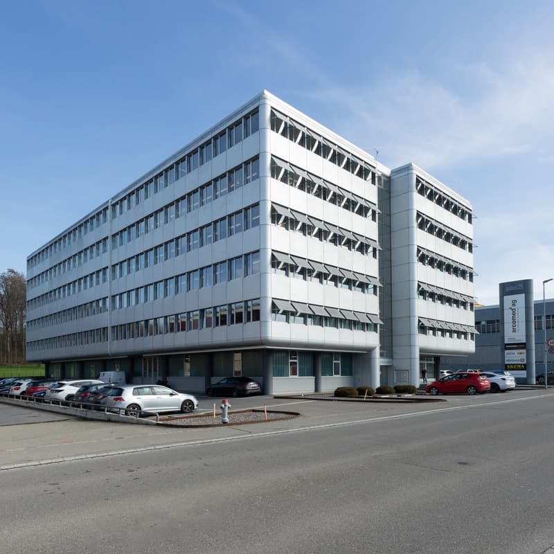 A modern, white office building with several floors, glass windows, and a paved parking area with multiple cars parked.