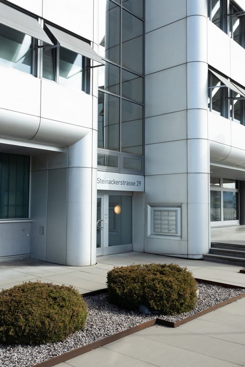 modern office building with sleek silver and glass exterior, Steinackerstrasse 29 address sign, double doors, windows with retractable awnings, circular security light above the entrance, and a small landscaped area in front