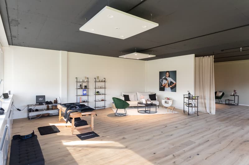 Open-concept studio apartment with hardwood floors, white walls, and modern furniture. The space includes a living area with a sofa and coffee table, a work area with a desk and shelving, and a massage table. The room is well-lit with recessed lighting an
