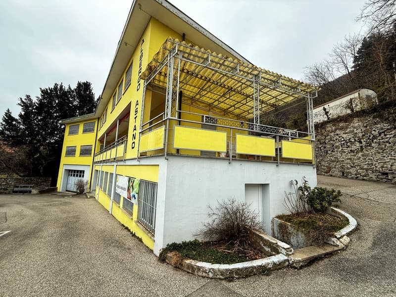 A two-story yellow building with a metal roof structure and balconies. The building has large windows and appears to be a commercial or community space. There is a small garden area in front of the building.