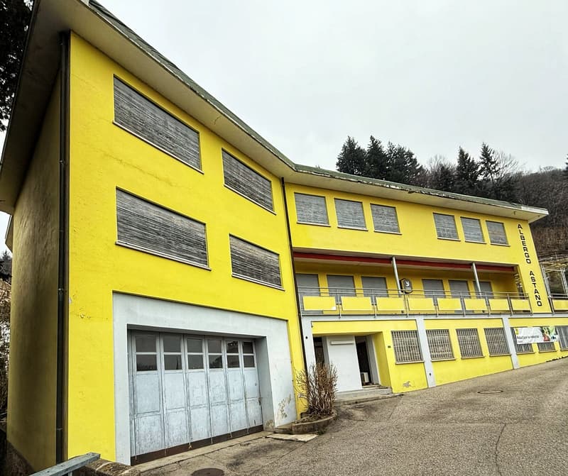 Two-story yellow building with a garage door and several windows. The building appears to be a commercial or industrial structure located in a forested area.