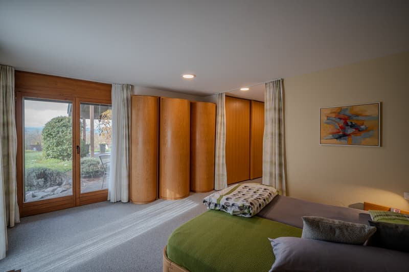 bedroom with large windows, wooden wardrobes, carpeted floor, bed with green blanket, pillows, painting on wall