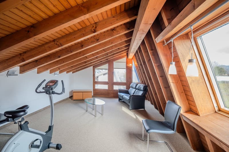 A room with wooden beams, a black leather couch, a glass table, a black chair, a wooden cabinet, a treadmill, and a window with a view of the outside.