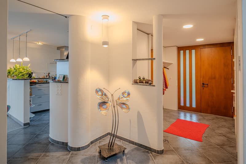 Entrance hall with white walls, gray floor, butterfly sculpture, wooden door, red rug, kitchen area with plants, cabinets, and counter