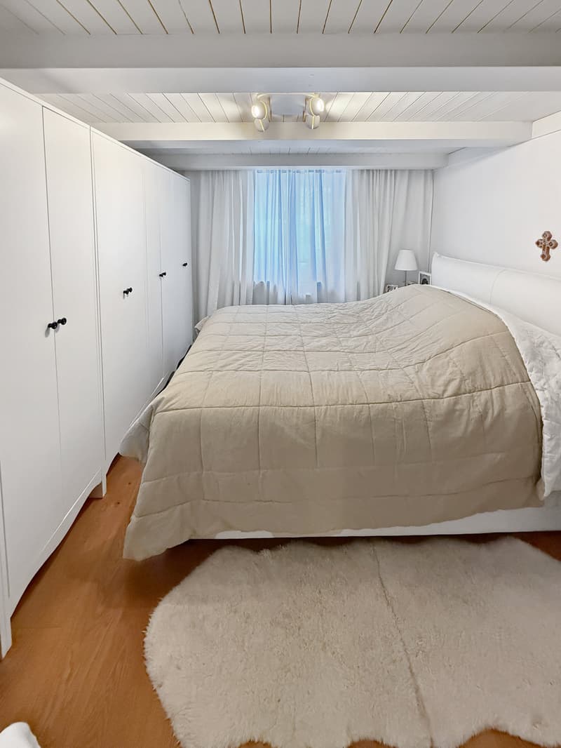 Bedroom with a bed, white walls, wooden floor, white cabinets, white rug, white curtains, lamp