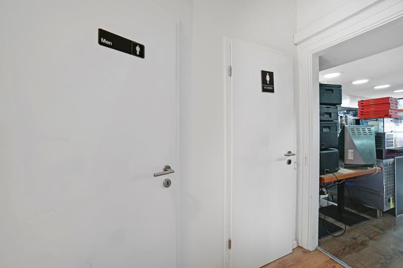 white walls, white door, silver door handle, black sign for men's restroom, black sign for women's restroom, open door leading to another room with a desk and equipment