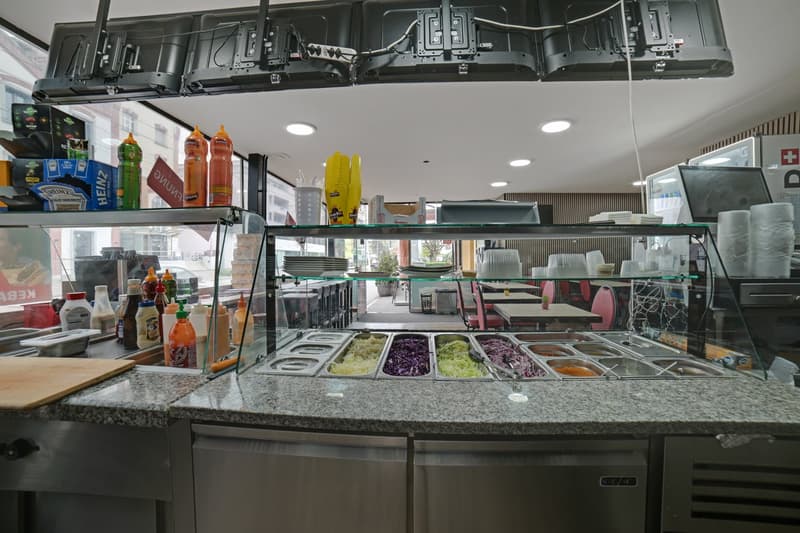 A restaurant kitchen with a counter, a glass display case with various food items, and a refrigerator. There are bottles, plates, and other items on the counter. The ceiling has lights and a surveillance camera. There are tables and chairs in the dining a