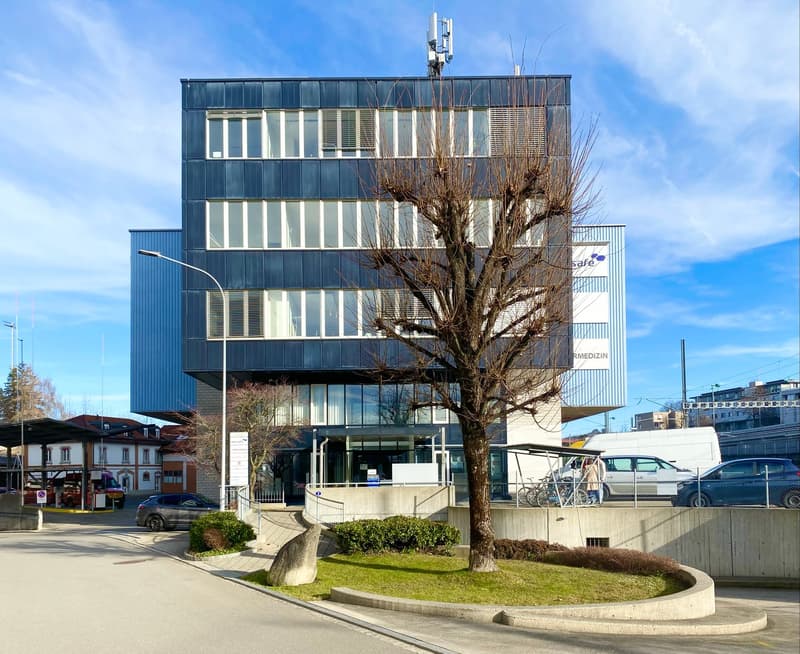 Office building with blue walls, white windows, a fenced entrance with bike racks, a street light, a tree, and cars parked in front