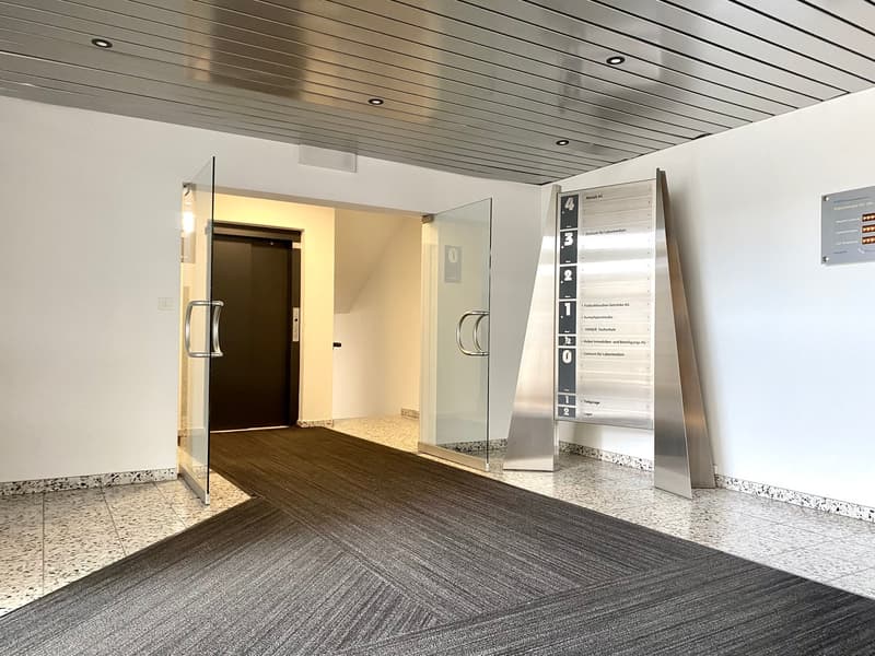 The image shows the entrance of a building with a dark wooden door, glass panels on either side, and a directory board displaying room numbers and names. The floor is covered with a gray patterned carpet, and the ceiling has a metal panel design.