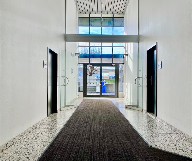 The image shows the entrance of a commercial building. The entrance has a glass door with a blue logo visible through it. The floor is made of terrazzo tiles, and the walls are white. The ceiling has a metal grid pattern. The space appears to be a lobby o