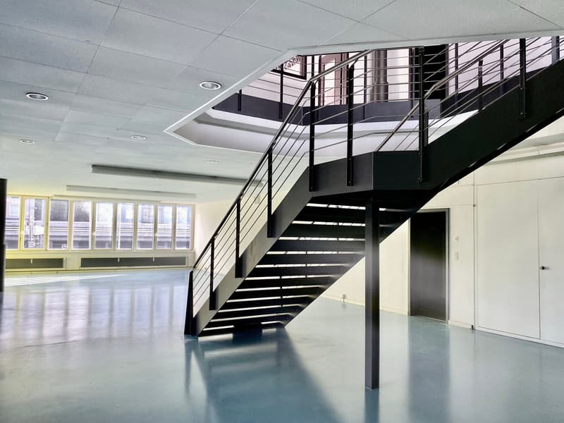 Empty room with a staircase, white walls, gray floor, large windows, metal railings
