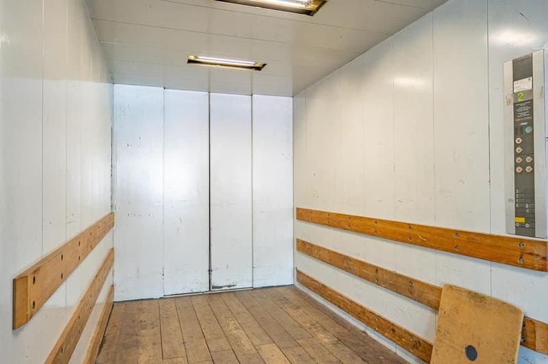 Empty room with white walls, wooden floor, two white doors, wooden panels on the wall, and an elevator control panel.