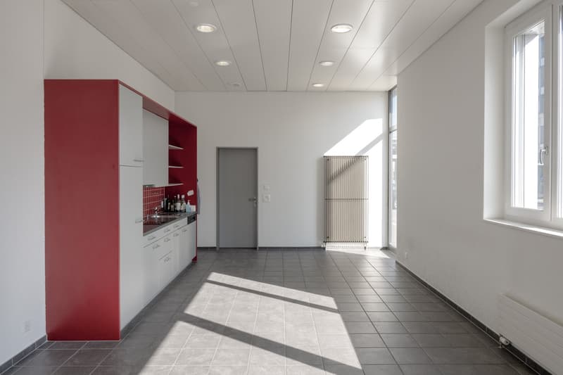 Empty room, tile flooring, white walls, red kitchen cabinets, stainless steel sink, glass window, electrical outlets, and heating radiator