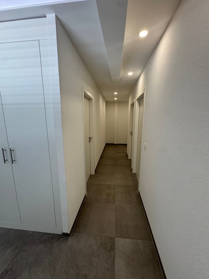 The image shows a long, narrow hallway with gray tile flooring and white walls. There are several doors leading off the hallway, and recessed lighting fixtures in the ceiling provide illumination.