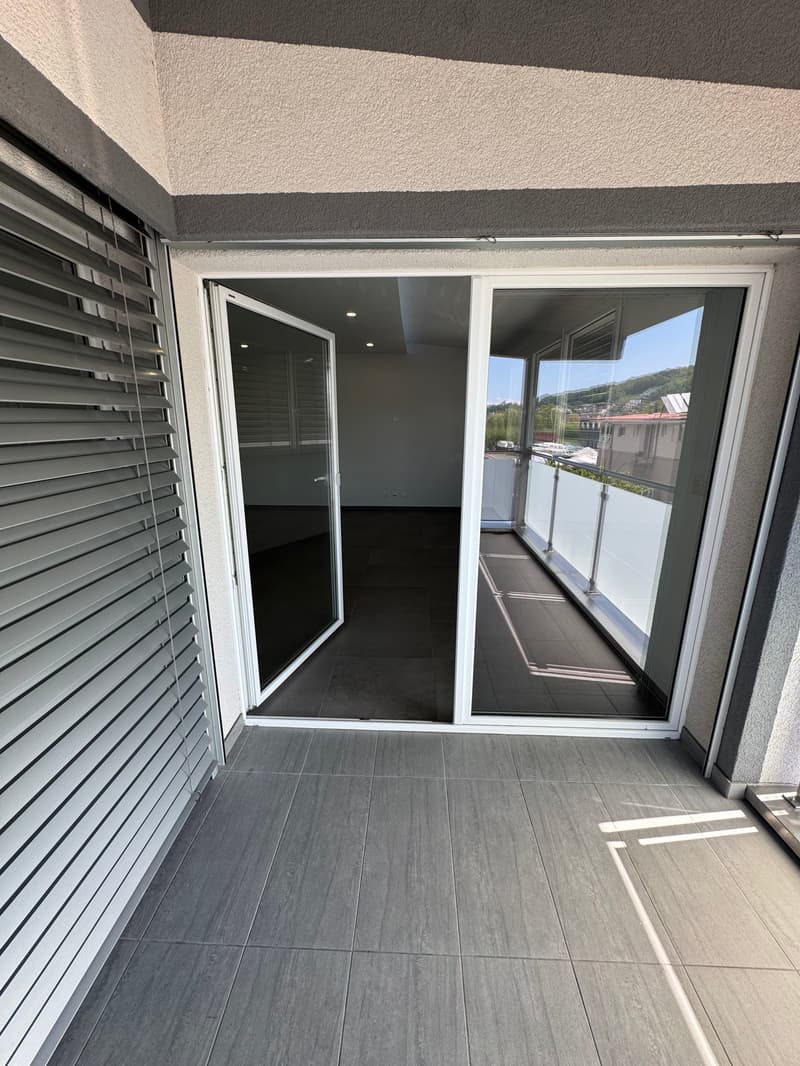 The image shows the interior of an apartment unit. It has a sliding glass door leading to a balcony or terrace, with a view of the surrounding area visible through the windows. The floor is covered in gray tile, and the walls are painted in a light color.